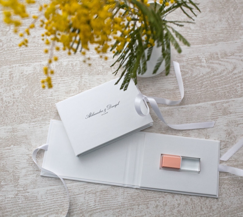 May include: White gift box with a white ribbon and a small pink and clear USB drive inside. The box has the text "Alexandra & Daniel" printed on the top.