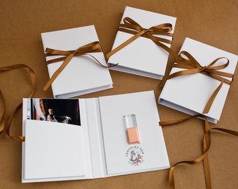 May include: White photo albums tied with brown satin ribbons. One album is open, revealing a photograph and a USB drive. The open album has the text "FRECKLED FOX" and a logo printed on the cover.