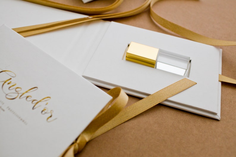 K&ouml;nnte beinhalten: Eine wei&szlig;e Box mit einem goldenen Band und einem goldenen und klaren Kristall-USB-Stick im Inneren. Die Box tr&auml;gt den Text "Angled'or" in goldenen Buchstaben.
