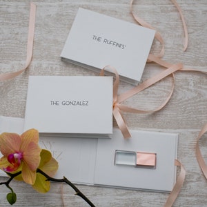 May include: Two white boxes with pink ribbon tied around them. The boxes are labeled "The Ruffin's" and "The Gonzalez". A white box with a clear and rose gold USB drive inside.