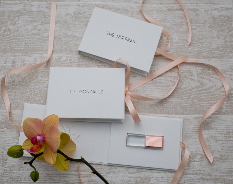 May include: Two white boxes with pink ribbon tied around them. The boxes are labeled "The Ruffin's" and "The Gonzalez". A white box with a clear and rose gold USB drive inside.
