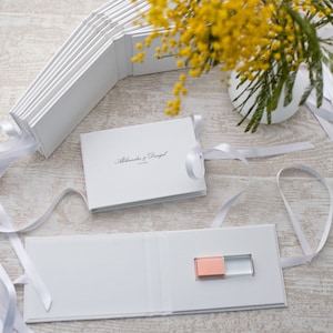 May include: White wedding photo album with a white ribbon and a small pink and clear USB drive inside. The album cover has the text "Aleksandra & Daniel" printed on it.