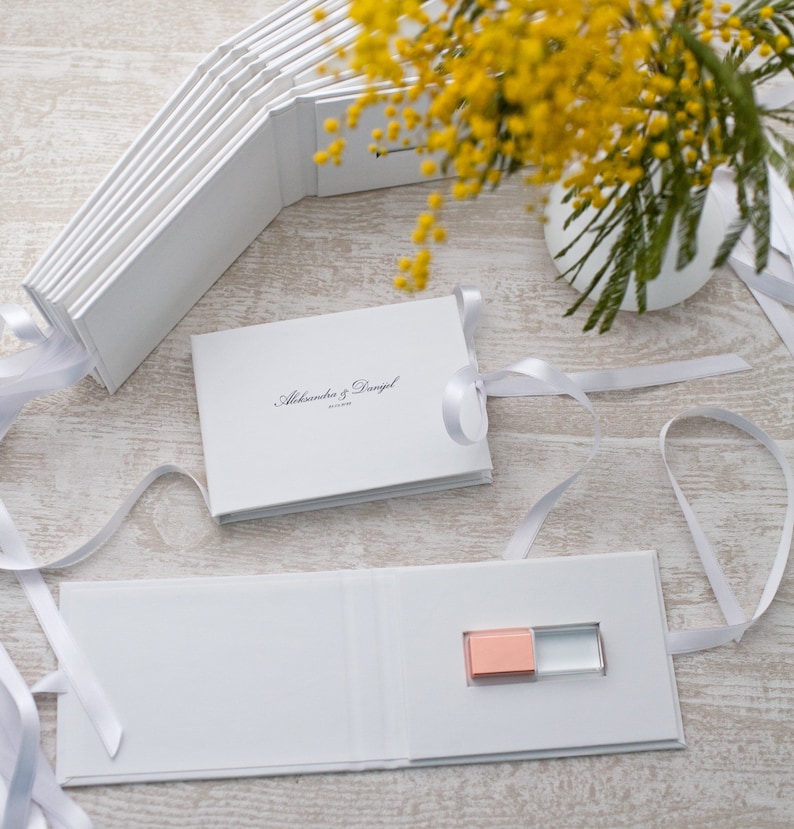 May include: White wedding photo album with a white ribbon and a small pink and clear USB drive inside. The album cover has the text "Aleksandra & Daniel" printed on it.