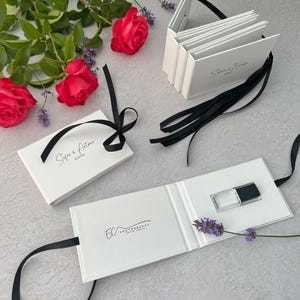 May include: Three white rectangular presentation folders with black ribbon ties. One folder is open, revealing a small clear glass USB drive with a black cap and the text EC PHOTOGRAPHY. The others feature the names Sofia & Antonio and Silvia & Nicola.
