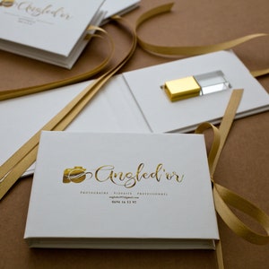 K&ouml;nnte beinhalten: Wei&szlig;e quadratische Boxen mit goldenen B&auml;ndern und einem goldenen und klaren USB-Stick. Die Box tr&auml;gt den Text "Angled'or" in goldenen Buchstaben und die Worte "Fotografie, Videografie, Professionell" in kleineren goldenen Buchstaben.