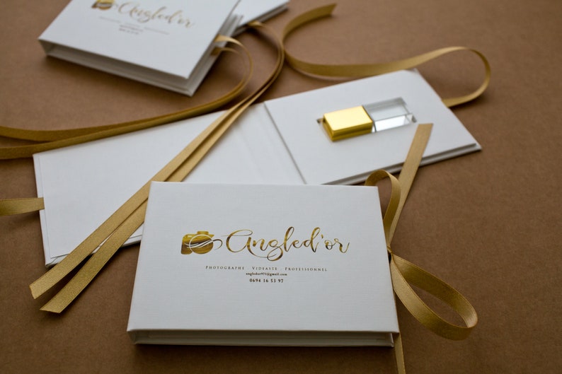 K&ouml;nnte beinhalten: Wei&szlig;e quadratische Boxen mit goldenen B&auml;ndern und einem goldenen und klaren USB-Stick. Die Box tr&auml;gt den Text "Angled'or" in goldenen Buchstaben und die Worte "Fotografie, Videografie, Professionell" in kleineren goldenen Buchstaben.