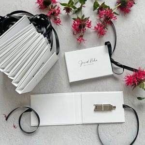 May include: White presentation boxes with black ribbon and the text "Jared Keha Photography". One box is open, revealing a silver USB drive. Pink flowers and greenery are arranged around the boxes.
