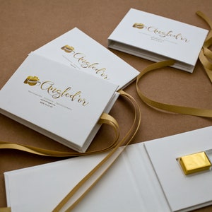 K&ouml;nnte beinhalten: Wei&szlig;e Boxen mit goldenem Band und einem goldenen und klaren Glas-USB-Stick. Die Boxen haben ein goldenes Kameralogo und den Text "Angled'or" auf der Vorderseite.