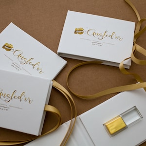 K&ouml;nnte beinhalten: Vier wei&szlig;e Boxen mit goldener Beschriftung und einem goldenen Kamerasymbol. Die Boxen sind mit "Angled'or Fotografie, K&uuml;nstlerisch, Professionell" beschriftet. Eine Box ist ge&ouml;ffnet und enth&auml;lt einen goldenen und transparenten USB-Stick.