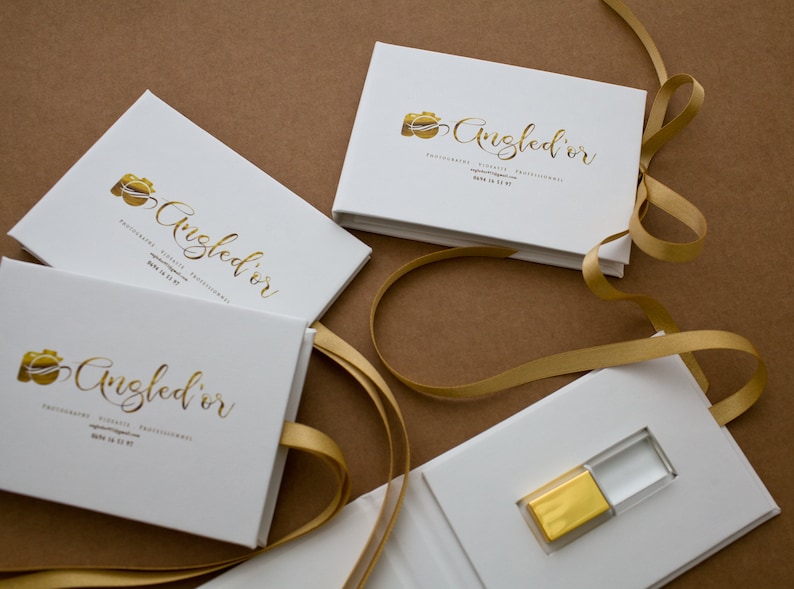 K&ouml;nnte beinhalten: Vier wei&szlig;e Boxen mit goldener Beschriftung und einem goldenen Kamerasymbol. Die Boxen sind mit "Angled'or Fotografie, K&uuml;nstlerisch, Professionell" beschriftet. Eine Box ist ge&ouml;ffnet und enth&auml;lt einen goldenen und transparenten USB-Stick.