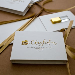 K&ouml;nnte beinhalten: Wei&szlig;e Box mit einem goldenen Band und einem goldenen Kameralogo mit dem Text "Angled'or" in goldener Schrift. Der Text unter dem Logo lautet "Photographie Vid&eacute;o Artistique Professionnels" und "angledor.wixsite.com". Die Telefonnummer "0694 16 11 97" ist ebenfalls enthalten.