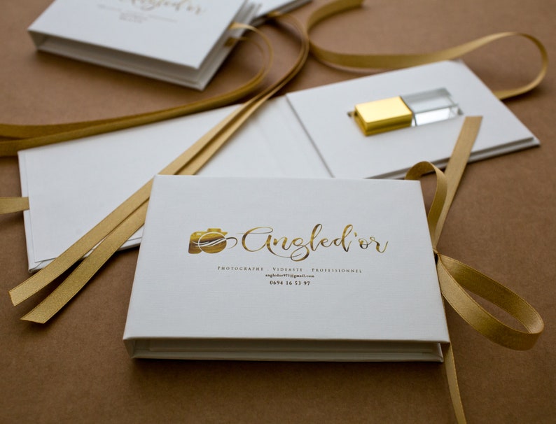 K&ouml;nnte beinhalten: Wei&szlig;e Box mit einem goldenen Band und einem goldenen Kameralogo mit dem Text "Angled'or" in goldener Schrift. Der Text unter dem Logo lautet "Photographie Vid&eacute;o Artistique Professionnels" und "angledor.wixsite.com". Die Telefonnummer "0694 16 11 97" ist ebenfalls enthalten.