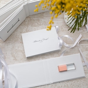 May include: White wedding album with a USB drive inside. The album is closed and has a white ribbon tied around it. The album has the text "Alexandra & Daniel" printed on the front.