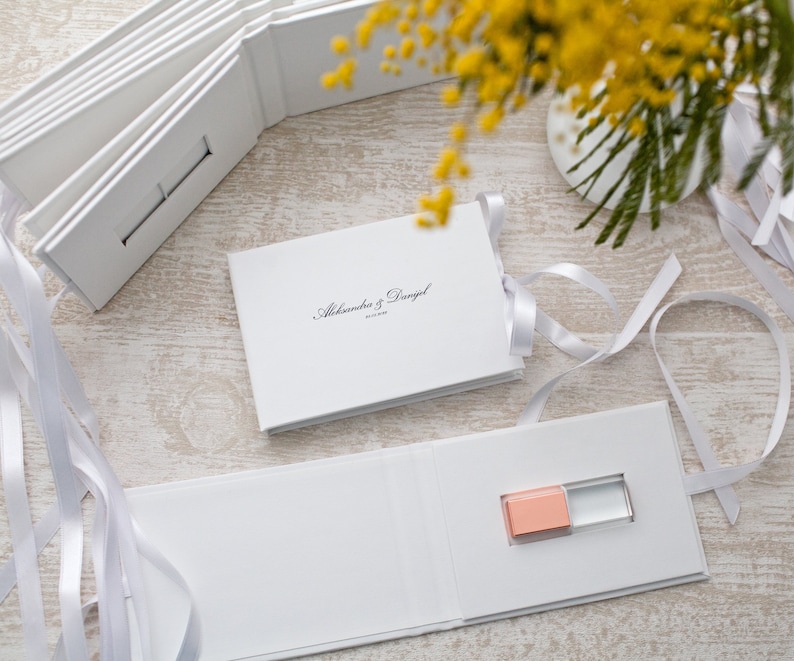 May include: White wedding album with a USB drive inside. The album is closed and has a white ribbon tied around it. The album has the text "Alexandra & Daniel" printed on the front.