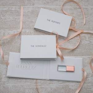 May include: White gift boxes with pink ribbon bows. The boxes are labeled "The Ruffin's", "The Gonzalez", and "Jenny Rodriguez". The box labeled "Jenny Rodriguez" is open and contains a small, clear USB drive with a rose gold cap.