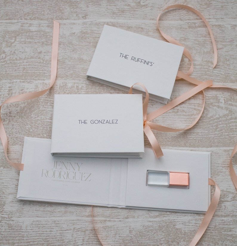 May include: White gift boxes with pink ribbon bows. The boxes are labeled "The Ruffin's", "The Gonzalez", and "Jenny Rodriguez". The box labeled "Jenny Rodriguez" is open and contains a small, clear USB drive with a rose gold cap.