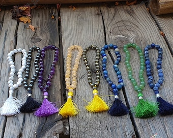 May include: Six beaded necklaces with tassels, each featuring a different color and type of stone. The necklaces are arranged in a row on a wooden surface.