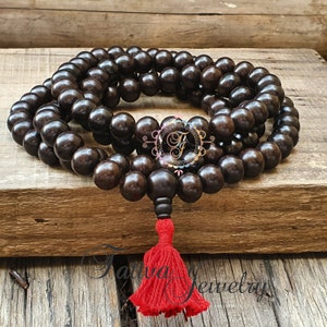 May include: A dark brown wooden mala bead necklace with a red tassel. The mala beads are round and have a smooth finish. The tassel is made of a soft, red fabric.