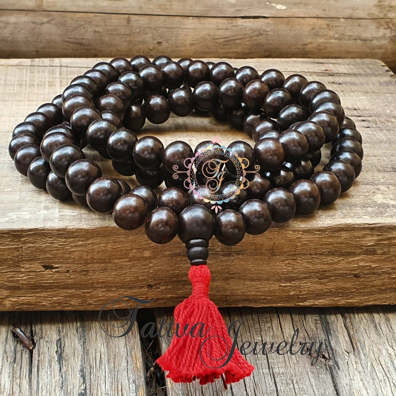 May include: A dark brown wooden mala bead necklace with a red tassel. The mala beads are round and have a smooth finish. The tassel is made of a soft, red fabric.
