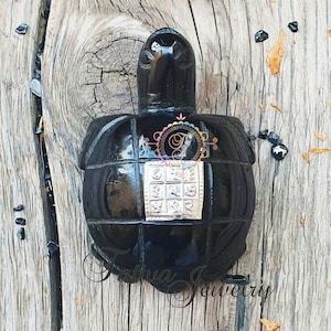 May include: A black, turtle-shaped decorative object with a silver square in the center. The object has a glossy finish and is set against a weathered wooden background. The text "Tattva Jewelry" is at the bottom.