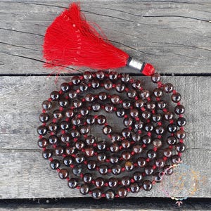 Puede incluir: Un collar mala con cuentas esféricas de color marrón oscuro ensartadas en un cordón rojo, dispuestas en un patrón de espiral circular. El collar termina con una borla roja vibrante sujeta por una tapa de metal plateado en la parte superior.