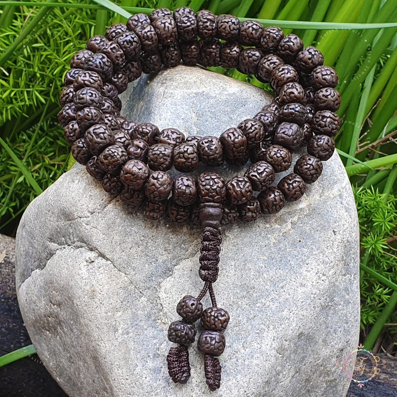 May include: A brown wooden mala bead necklace with a tassel. The beads are round and have a textured surface. The necklace is lying on a gray rock.