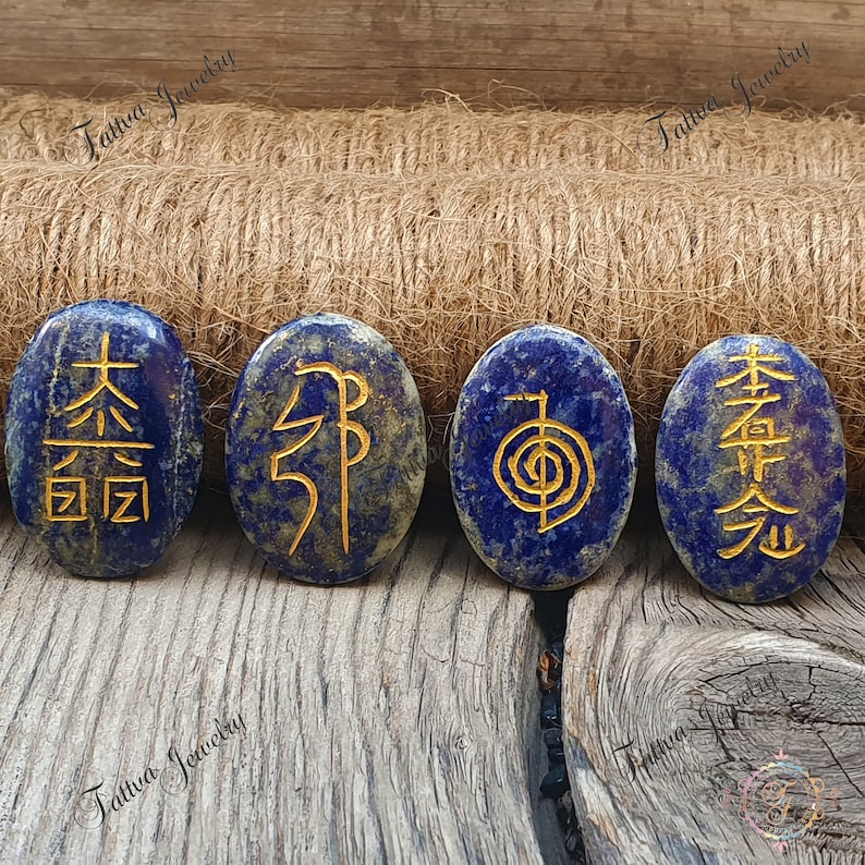 May include: Four oval-shaped lapis lazuli stones with gold symbols inscribed on them. The stones are arranged in a row on a wooden surface.