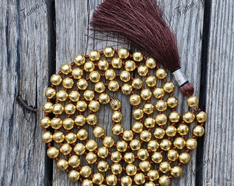 May include: A gold-colored mala bead necklace with a brown tassel. The beads are round and evenly spaced on a brown cord. The tassel is a deep brown color and is attached to the top of the necklace. The necklace is displayed on a weathered wooden surface.