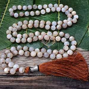 May include: A white moonstone mala with a brown tassel. The mala is made of round beads and is strung on a brown cord.