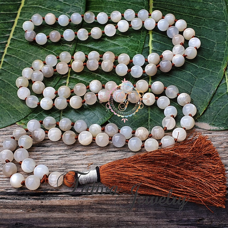 May include: A white moonstone mala with a brown tassel. The mala is made of round beads and is strung on a brown cord.