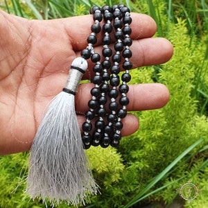 May include: A black beaded mala with a silver tassel. The mala is made of round beads and is strung on a silver cord.