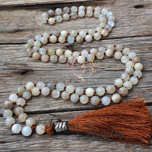 May include: A white moonstone mala necklace with a brown tassel. The necklace is made of 108 beads and is strung on a brown cord.