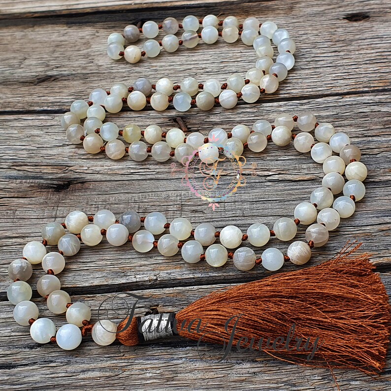 May include: A white moonstone mala necklace with a brown tassel. The necklace is made of 108 beads and is strung on a brown cord.
