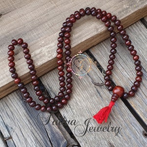 May include: A dark red wooden mala bead necklace with a red tassel. The mala has 108 beads and is approximately 18 inches long.