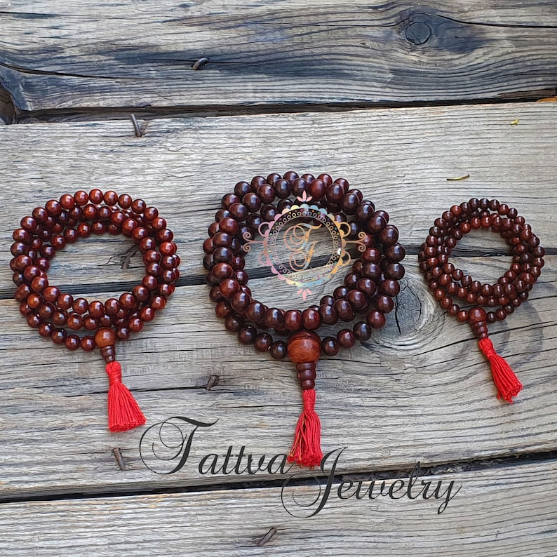 May include: Three wooden mala bead necklaces with red tassels. The necklaces are different sizes and are arranged in a row on a wooden surface.
