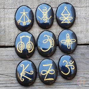May include: Nine black oval-shaped stones with gold symbols etched on them. The symbols include a cross, a spiral, a triangle, a circle, a crescent moon, a star, a letter Z, and two Om symbols.