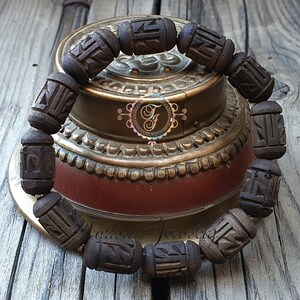 Pode incluir: Um bracelete de madeira castanho escuro com detalhes entalhados. O bracelete &eacute; feito de v&aacute;rias contas e est&aacute; sobre uma superf&iacute;cie de madeira.