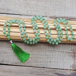 May include: A green jade mala bead necklace with a green tassel. The necklace is made of round beads and is strung on a thin cord. The tassel is attached to the end of the necklace.