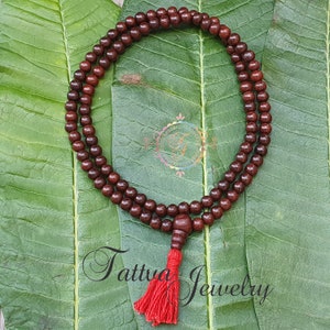May include: A brown wooden mala bead necklace with a red tassel. The necklace is laid out on a green leaf background.