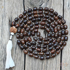 Può includere: Una collana mala fatta di semi marrone scuro infilati su un filo bianco. La collana ha un nappa decorativa e una perla di rudraksha. Le perle di forma ovale hanno un aspetto liscio e lucido. Lo sfondo è una superficie di legno invecchiato.