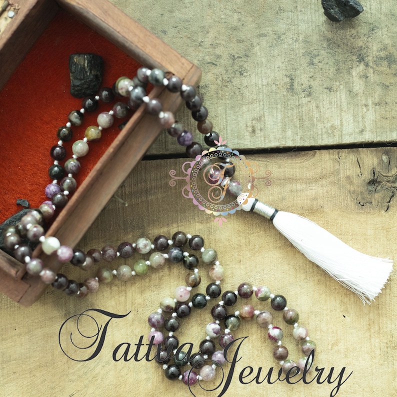 May include: A multi-colored gemstone mala necklace with a white tassel. The necklace is made of round beads and is displayed in a wooden box.
