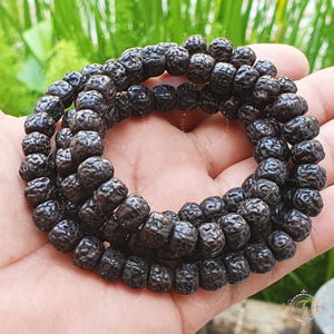 May include: A close-up of a dark brown beaded necklace made of round, textured beads. The necklace is wrapped around a hand, showing the length and detail of the beads.