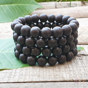 Puede incluir: Un conjunto de pulseras de cuentas de madera negra. Las cuentas son redondas y lisas, y las pulseras están apiladas unas sobre otras.