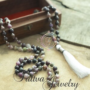 May include: A multi-colored gemstone mala necklace with a white tassel. The necklace is made of round beads and is strung on a silver cord. The necklace is displayed in a wooden box.