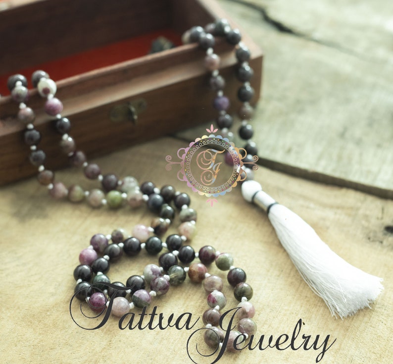 May include: A multi-colored gemstone mala necklace with a white tassel. The necklace is made of round beads and is strung on a silver cord. The necklace is displayed in a wooden box.