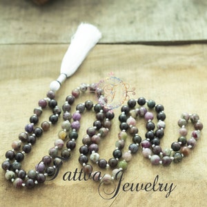 May include: A mala or prayer beads made of dark purple, green, and pink stones with a white tassel. The beads are strung on a white cord.