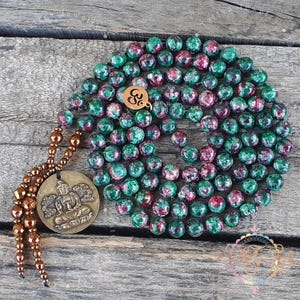 May include: A mala necklace featuring round green and magenta zoisite beads arranged in a circular pattern. The necklace includes a small round brass charm with an Om symbol and a larger round brass pendant depicting a seated figure.
