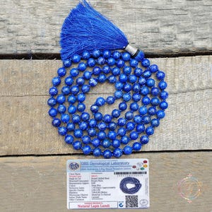 May include: A blue beaded mala necklace with a tassel. The necklace is made of round, deep blue beads. A tag at the bottom reads "Natural Lapis Lazuli".