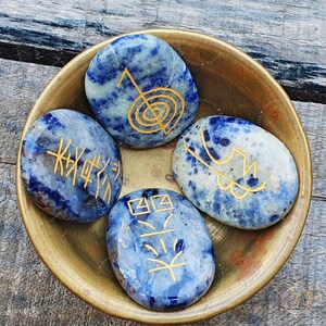 Puede incluir: Cuatro piedras lisas de forma ovalada, azules y blancas, con s&iacute;mbolos dorados grabados en ellas. Las piedras est&aacute;n en un cuenco de lat&oacute;n.