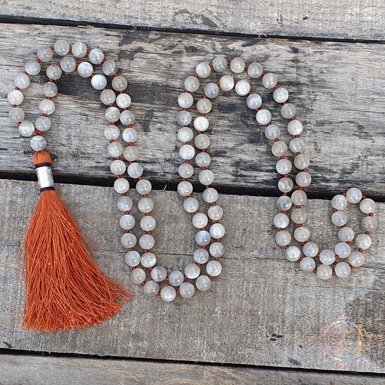 May include: A gray moonstone mala necklace with a tassel made of orange thread. The necklace is made of round beads and is strung on a thin cord.